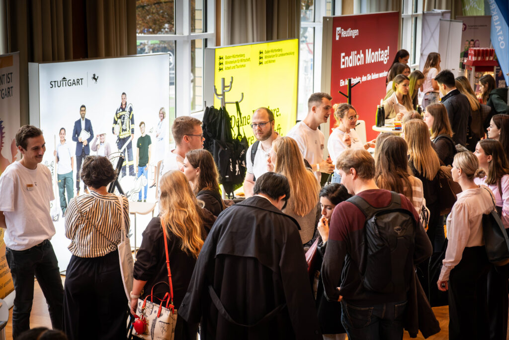 Menschenmenge an Messeständen; mit Postern in verschiedenen Farben und vielen jungen Menschen, die an den Ständen stehen und sich unterhalten.