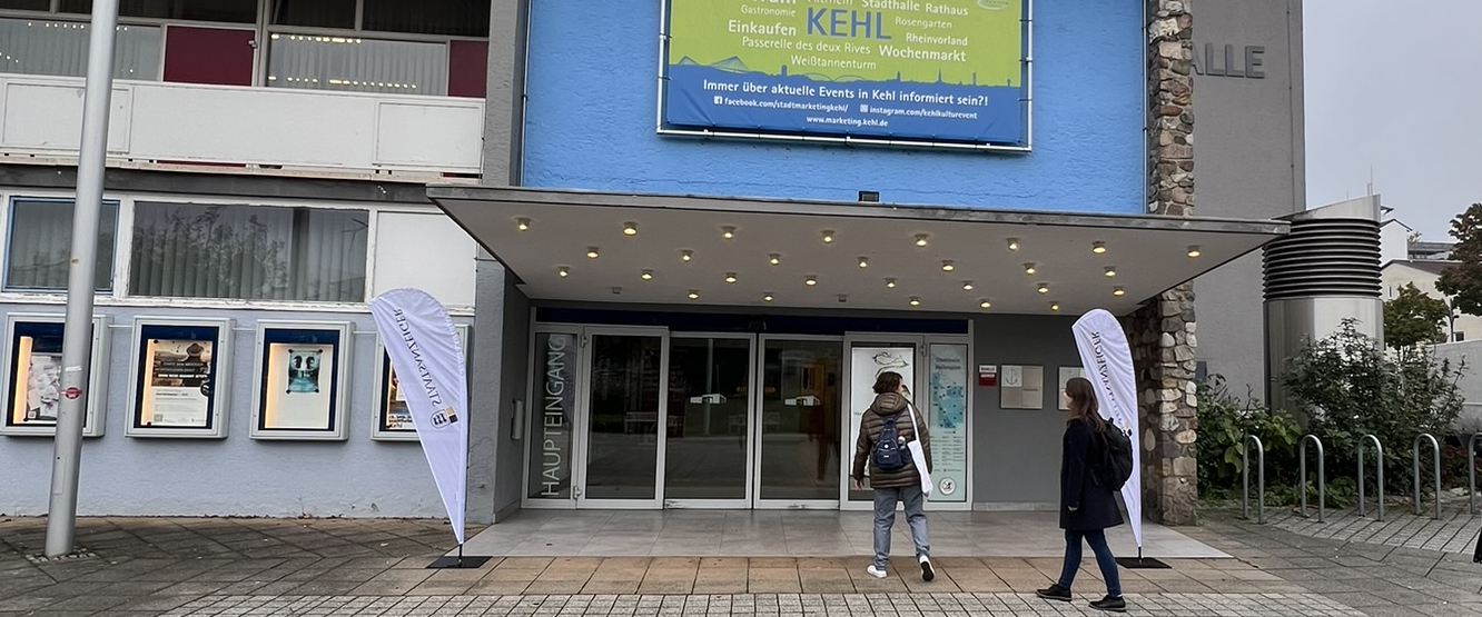 Eingang eines Gebäudes mit Vordach, zwei Personen davor; Fahnen links und rechts; Leuchttafel mit „KEHL“, Schild „HAUPTEINGANG“.