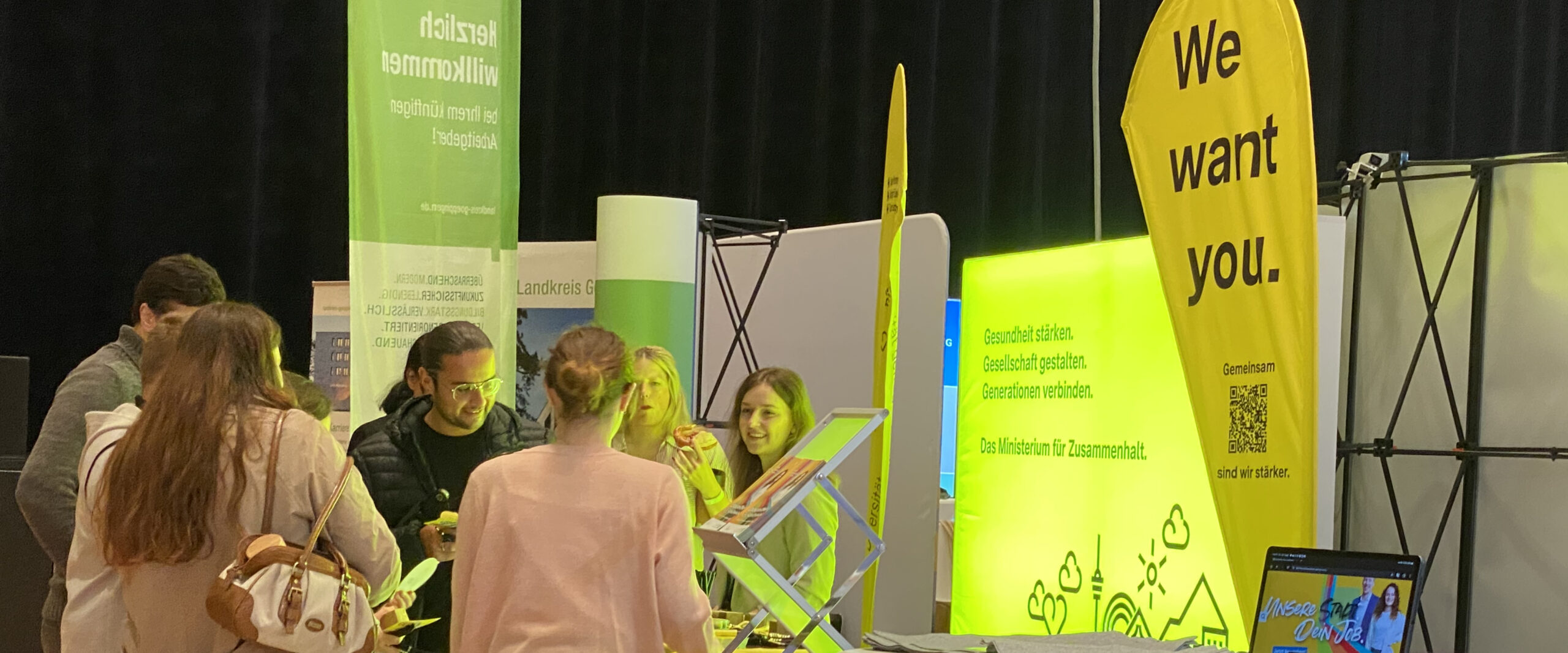 Personen an Messestand; gelbe Beachflag „We want you.“; Leuchtwand: „Gesundheit stärken. Gesellschaft gestalten. Generationen verbinden.“