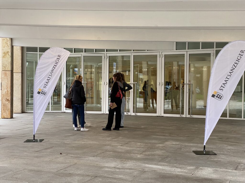 Drei Personen vor Glaseingang; zwei Beachflags mit Aufdruck „STAATSANZEIGER“ links und rechts.