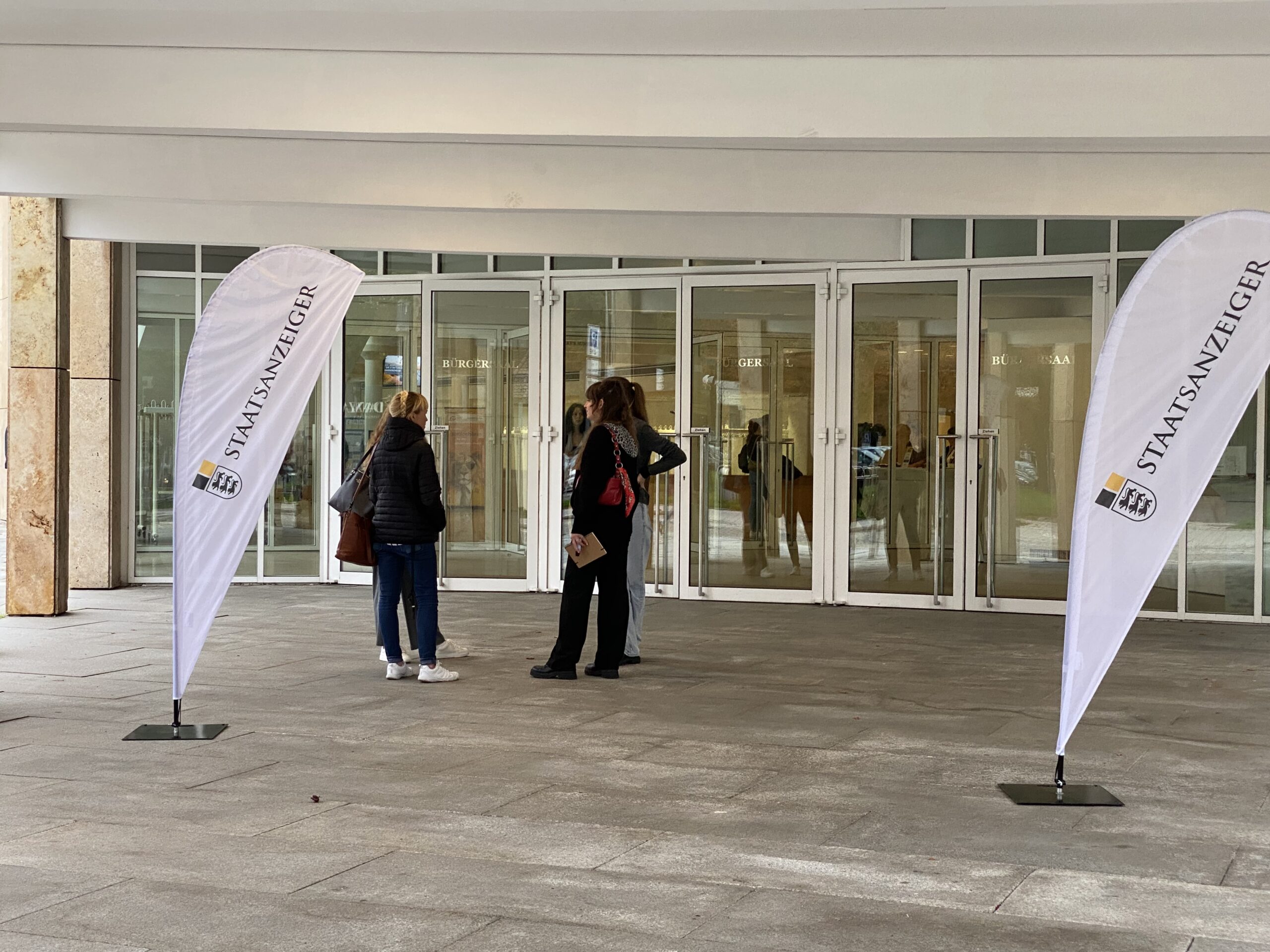 Drei Personen vor Glaseingang; zwei Beachflags mit Aufdruck „STAATSANZEIGER“ links und rechts.