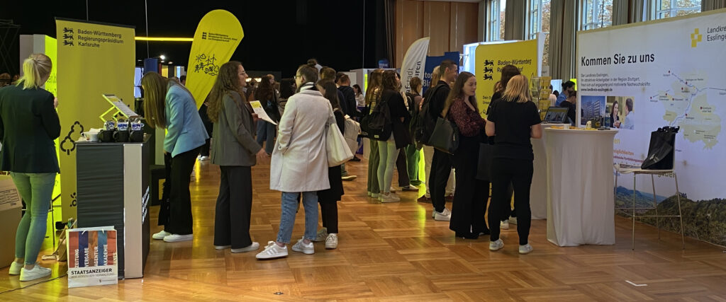 Messehalle mit Ständen und Besuchenden; Gelbe Schilder mit beispielsweise "Baden-Württemberg Regierungspräsidium Karlsruhe“ und „Landkreis Esslingen".
