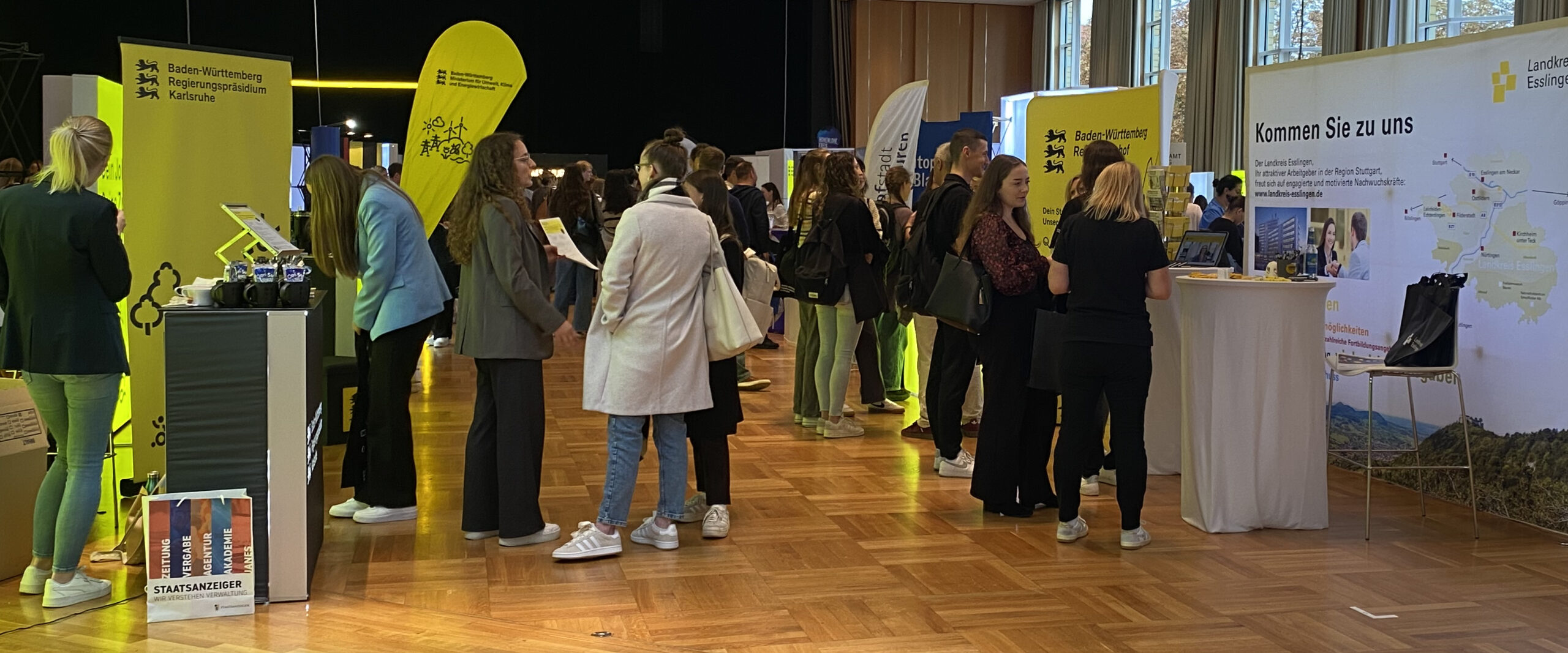 Messehalle mit Ständen und Besuchenden; Gelbe Schilder mit beispielsweise "Baden-Württemberg Regierungspräsidium Karlsruhe“ und „Landkreis Esslingen".