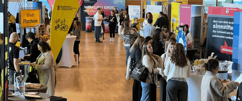 Messehalle mit Ständen und vielen Menschen; Banner mit „Stadt Pforzheim“, „Heidelberg“ und weiteren Logos.