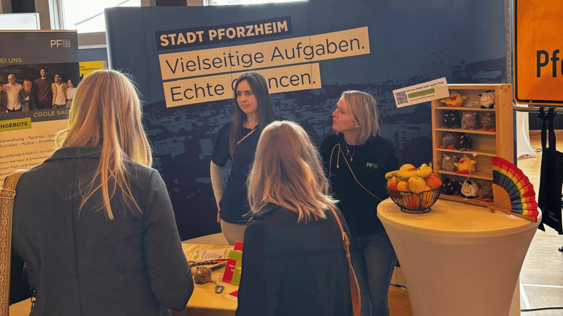 Messestand Stadt Pforzheim „Vielseitige Aufgaben. Echte Chancen.“ Drei Frauen im Gespräch, Tisch mit Obst und Stofftieren.