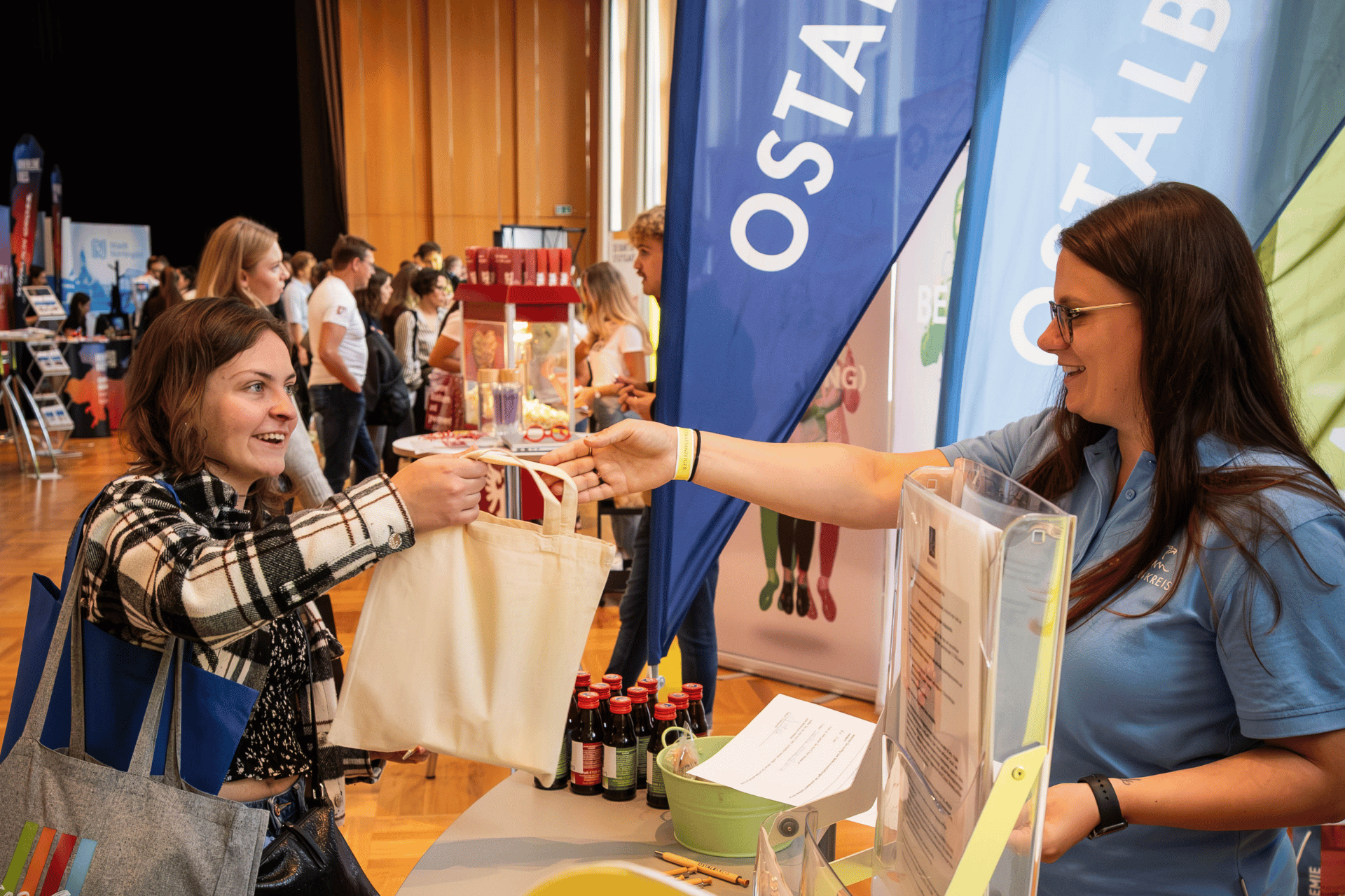 Zwei Frauen an Messestand, eine überreicht Stofftasche; auf Tisch stehen Flaschen und Flyer.