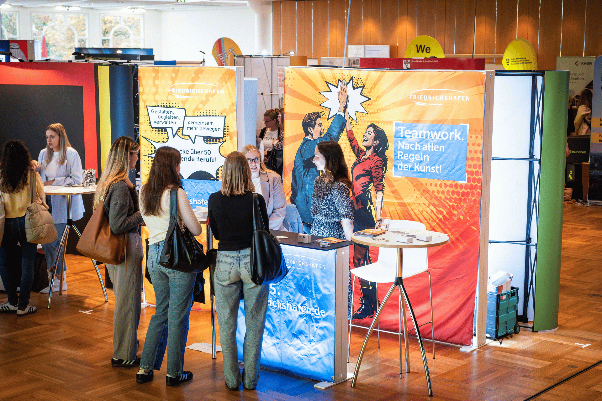 Messestand „Friedrichshafen“ mit Plakat „Teamwork. Nach allen Regeln der Kunst!“, mehrere Personen im Gespräch.