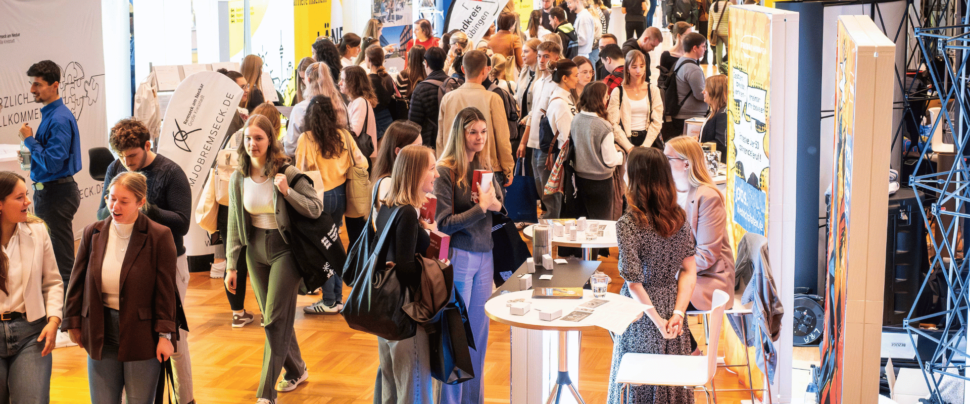 Große Messehalle mit dichtem Besucherandrang an Ständen, Menschen im Gespräch und an Stehtischen mit Flyern.