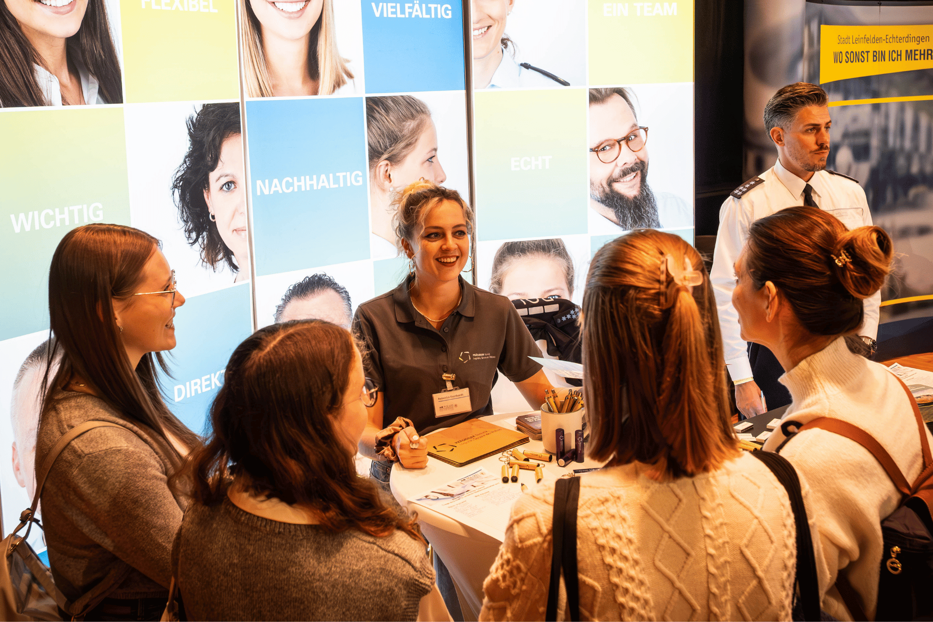 Fünf Personen stehen an einem Messestand im Gespräch; im Hintergrun Wörter wie „FLEXIBEL“, „NACHHALTIG“, „ECHT“.