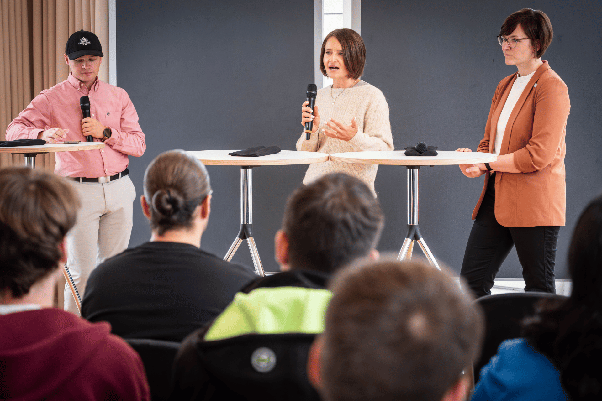 Drei Personen stehen mit Mikrofonen an Stehtischen vor Publikum und sprechen.