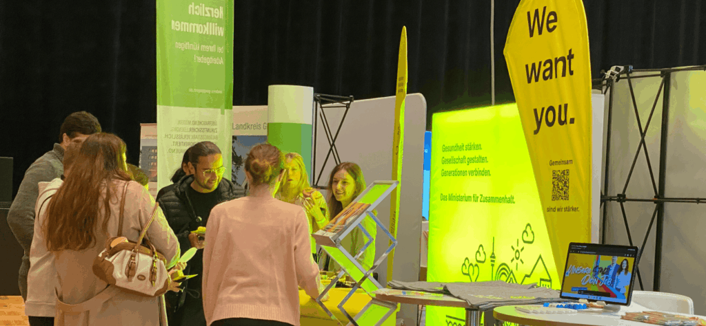 Personen an Messestand; gelbe Beachflag „We want you.“; Leuchtwand: „Gesundheit stärken. Gesellschaft gestalten. Generationen verbinden.“