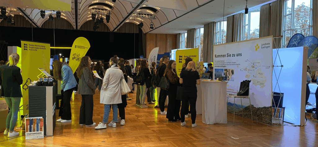 Besucherinnen und Besucher informieren sich an Messeständen auf einer Karrieremesse in einer hellen Veranstaltungshalle, unter anderem am Stand des Regierungspräsidiums Karlsruhe und des Landkreises Esslingen.
