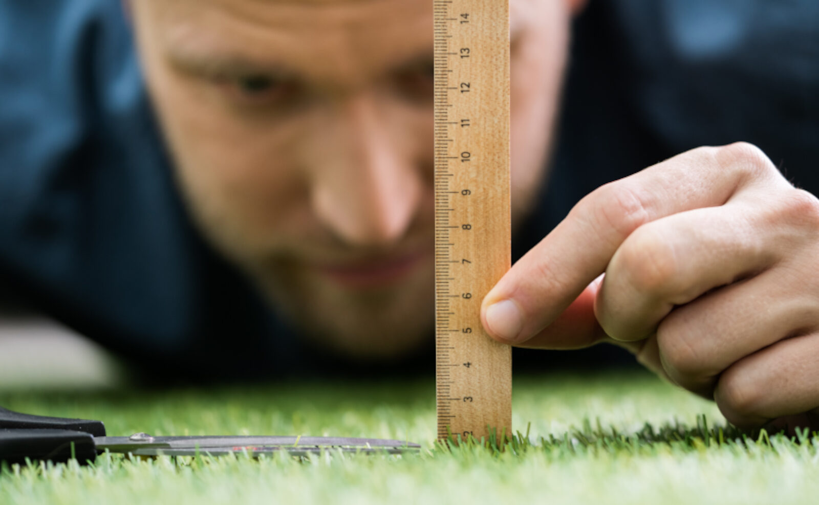 Ein Mann misst mit einem Holzlineal die Höhe eines Rasens; neben dem Lineal liegt eine Schere im Gras.